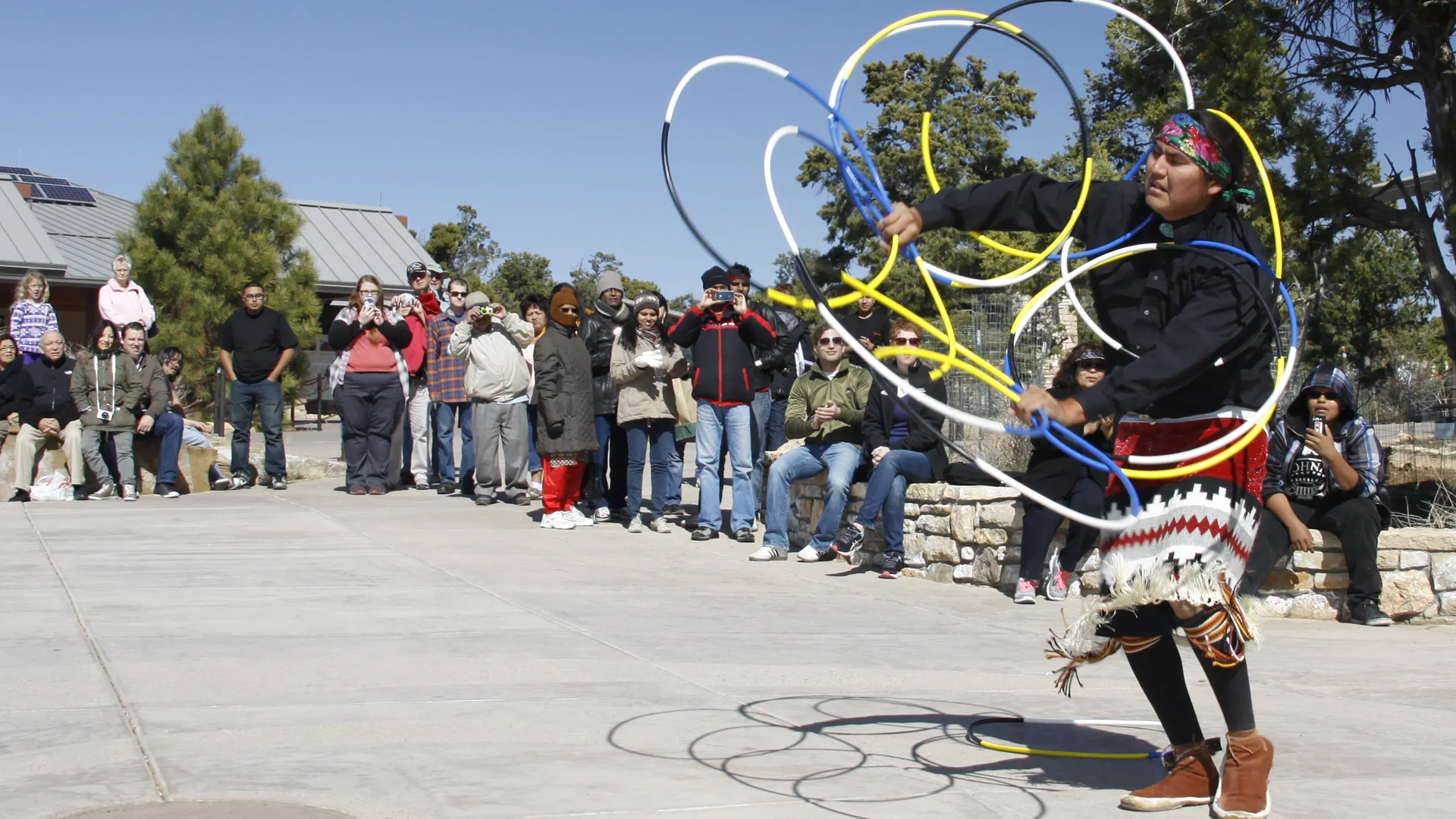 A hoop dancer in a woven-pattern skirt spins a cluster of coloured hoops around his body while visitors watch and take photographs.